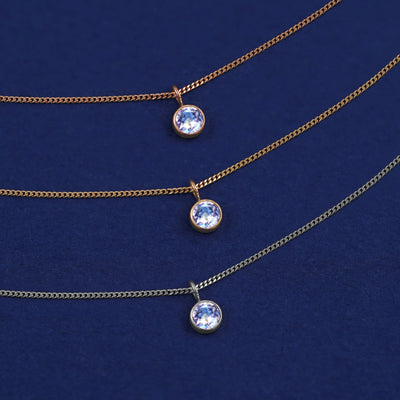 Three versions of the Aquamarine Necklace shown in options of yellow, white and rose gold on a dark blue background