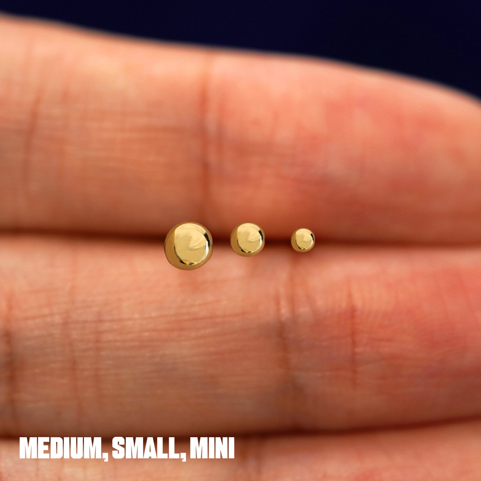 Solid 14k yellow gold Medium, Small, and Mini Ball Earrings in between a model's fingers