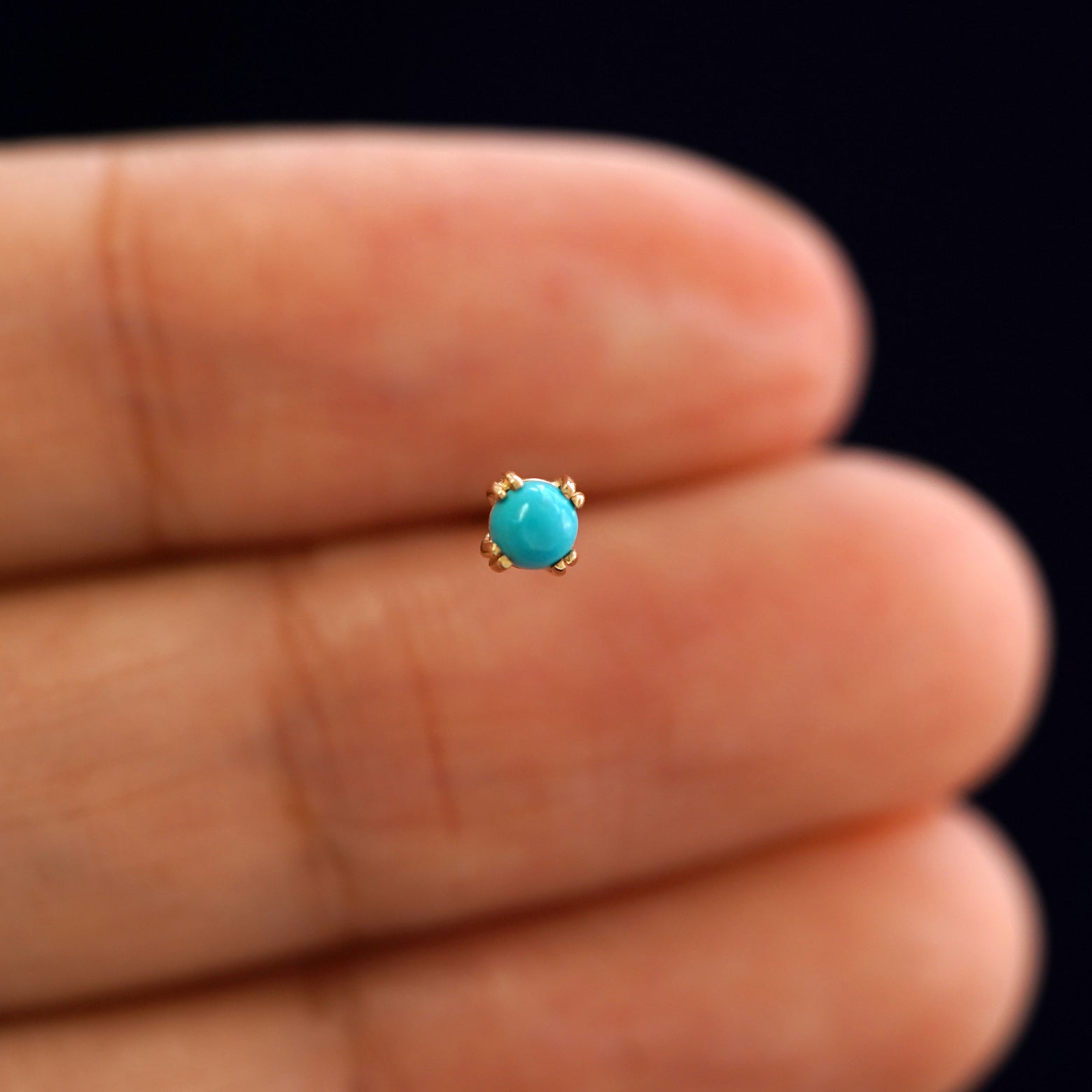 A model holding a single 3mm turquoise gemstone earring between their fingers to show the setting