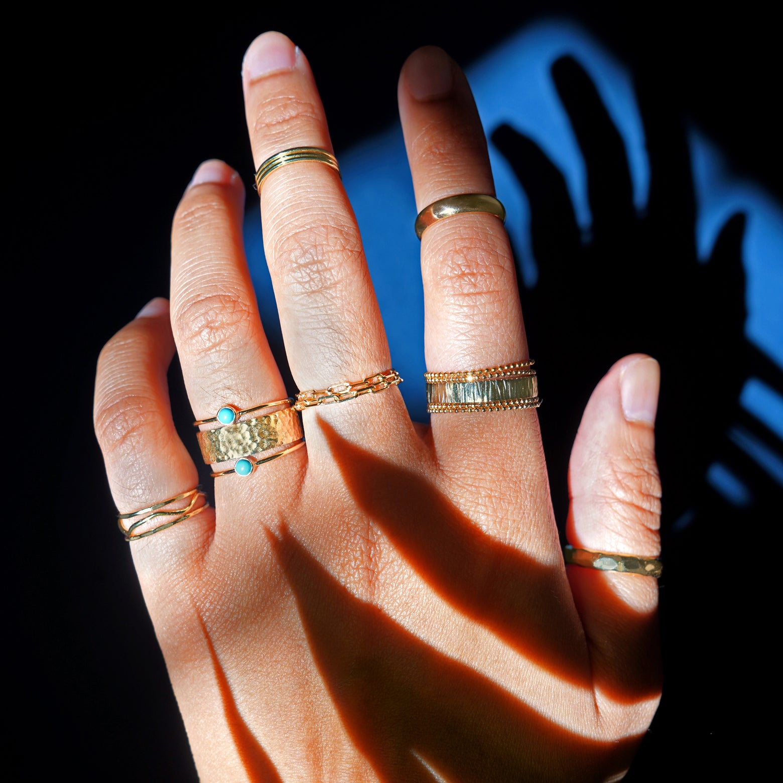 A model's hand lit by a strong light wearing stacks of different Automic Gold rings including two Turquoise Rings 