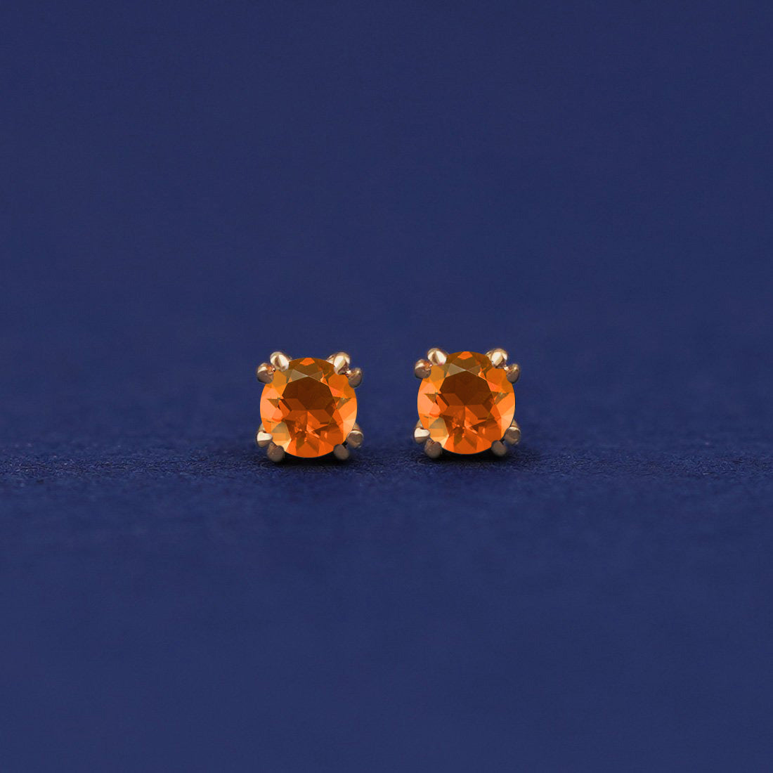 A pair of 14 karat gold stud earrings with 3 millimeter round orange Fire Opals on a dark blue background