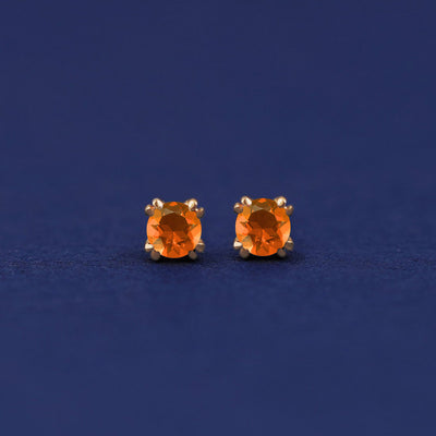 A pair of 14 karat gold stud earrings with 3 millimeter round orange Fire Opals on a dark blue background