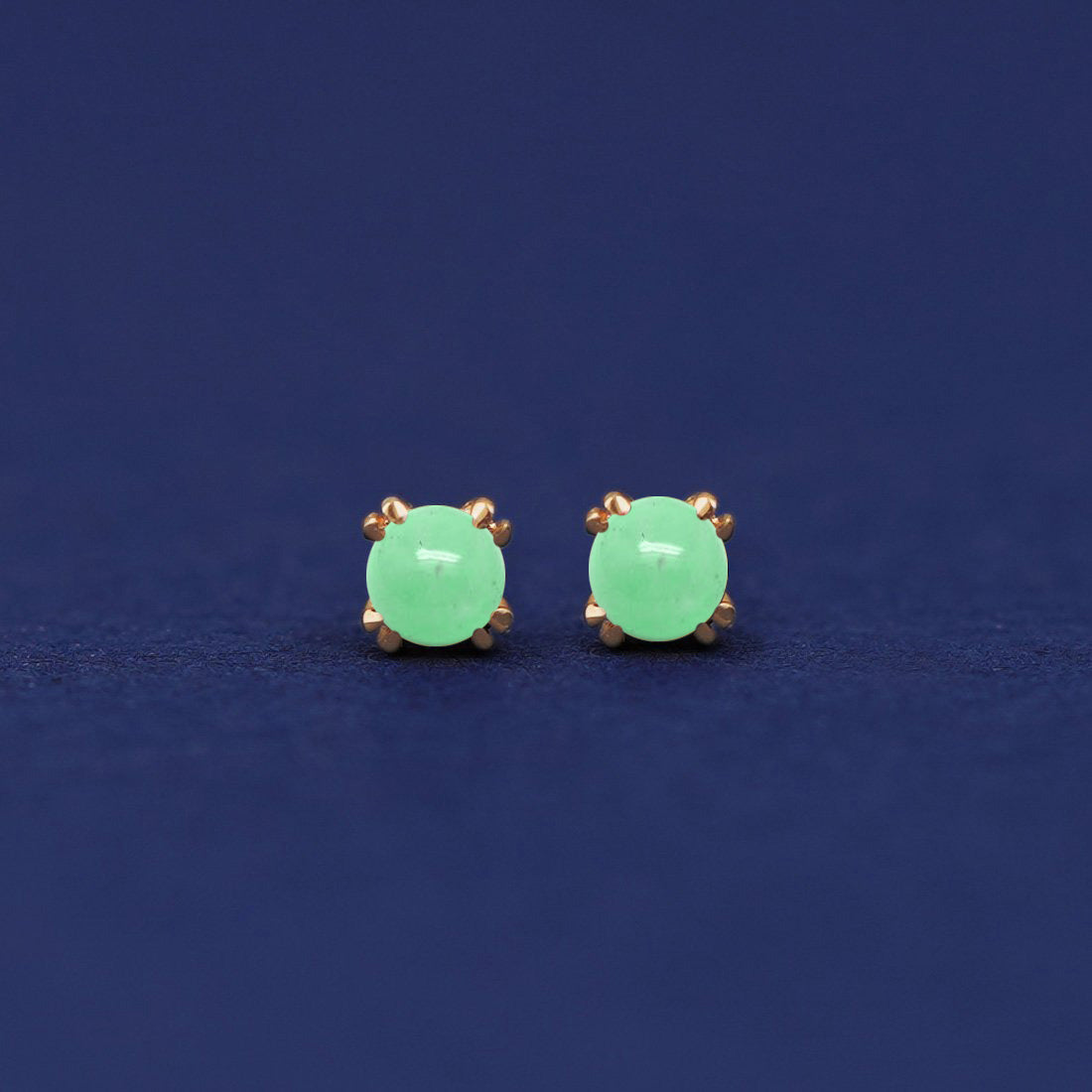 A pair of 14 karat gold stud earrings with 3 millimeter round Green Jades on a dark blue background