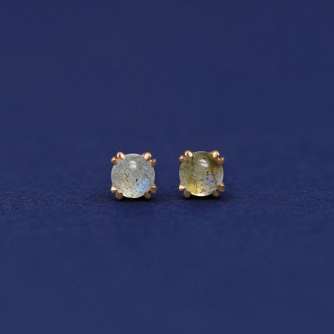 A pair of 14 karat gold stud earrings with 3 millimeter round Labradorites on a dark blue background