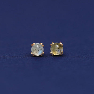 A pair of 14 karat gold stud earrings with 3 millimeter round Labradorites on a dark blue background