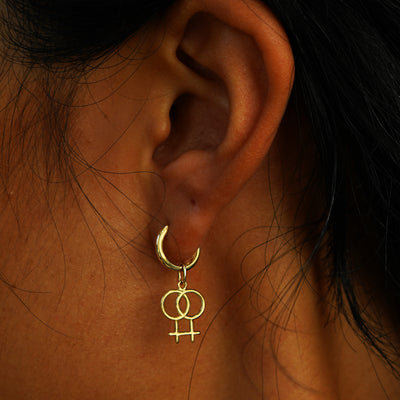 A model's ear wearing a yellow gold Lesbian Symbol Charm on a Curvy Huggie Hoop