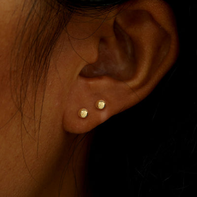 A model's ear wearing two 14k gold Medium Ball Flatback Piercings in two lobe piercings