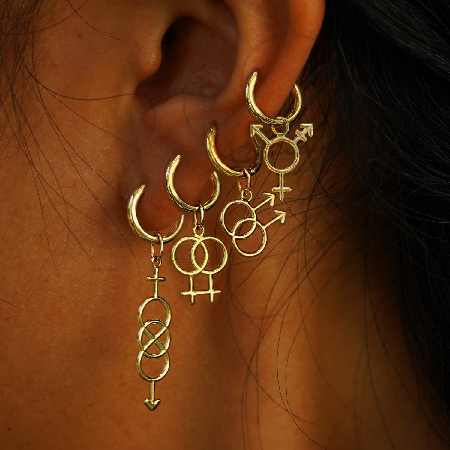 A model's ear wearing yellow gold Bisexual, Lesbian, Gay, and Transgender symbol charms on four Medium Curvy Huggies