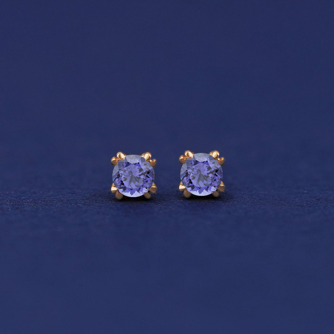 A pair of 14 karat gold studs earrings with 3 millimeter round Tanzanite gemstones on a dark blue background