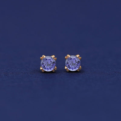 A pair of 14 karat gold studs earrings with 3 millimeter round Tanzanite gemstones on a dark blue background