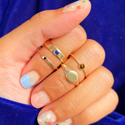 A hand gripping a blue sweater and wearing a gemstone signet, tiger eye, sapphire industrial, and black diamond bar rings