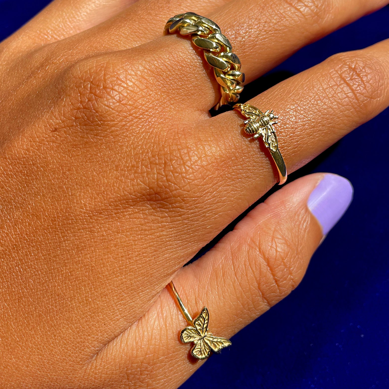 A hand with a Butterfly ring on the thumb, Curvy Bee Ring on the pointer finger, and a Miami Cuban Ring on the middle finger