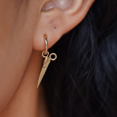 Close up view of a model's ear wearing a yellow gold Scissors Charm on a Curvy Huggie Hoop showing the scissors closed