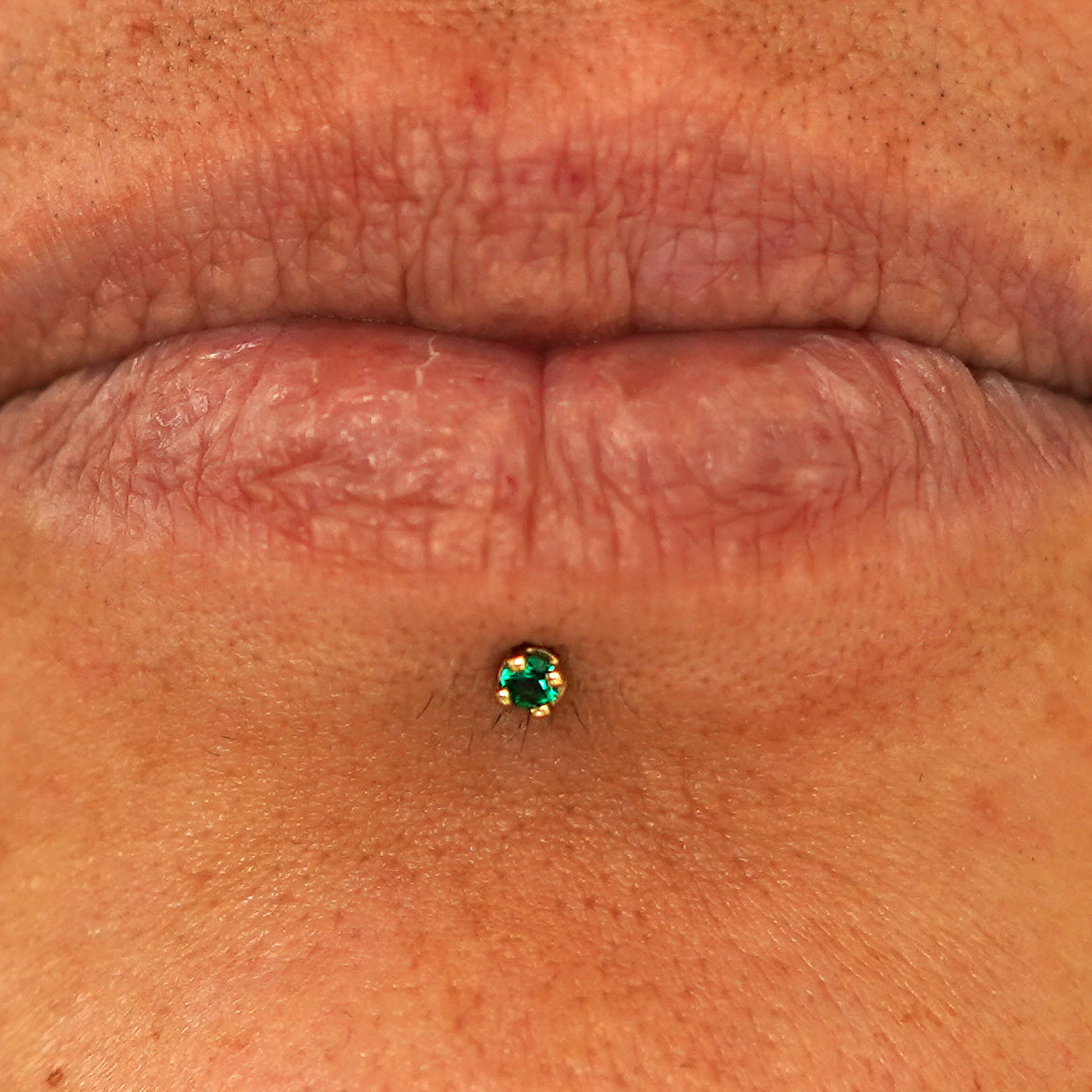 Close up view of a model's lips wearing a 14k yellow gold 2mm emerald gemstone Flatback in a labret piercing