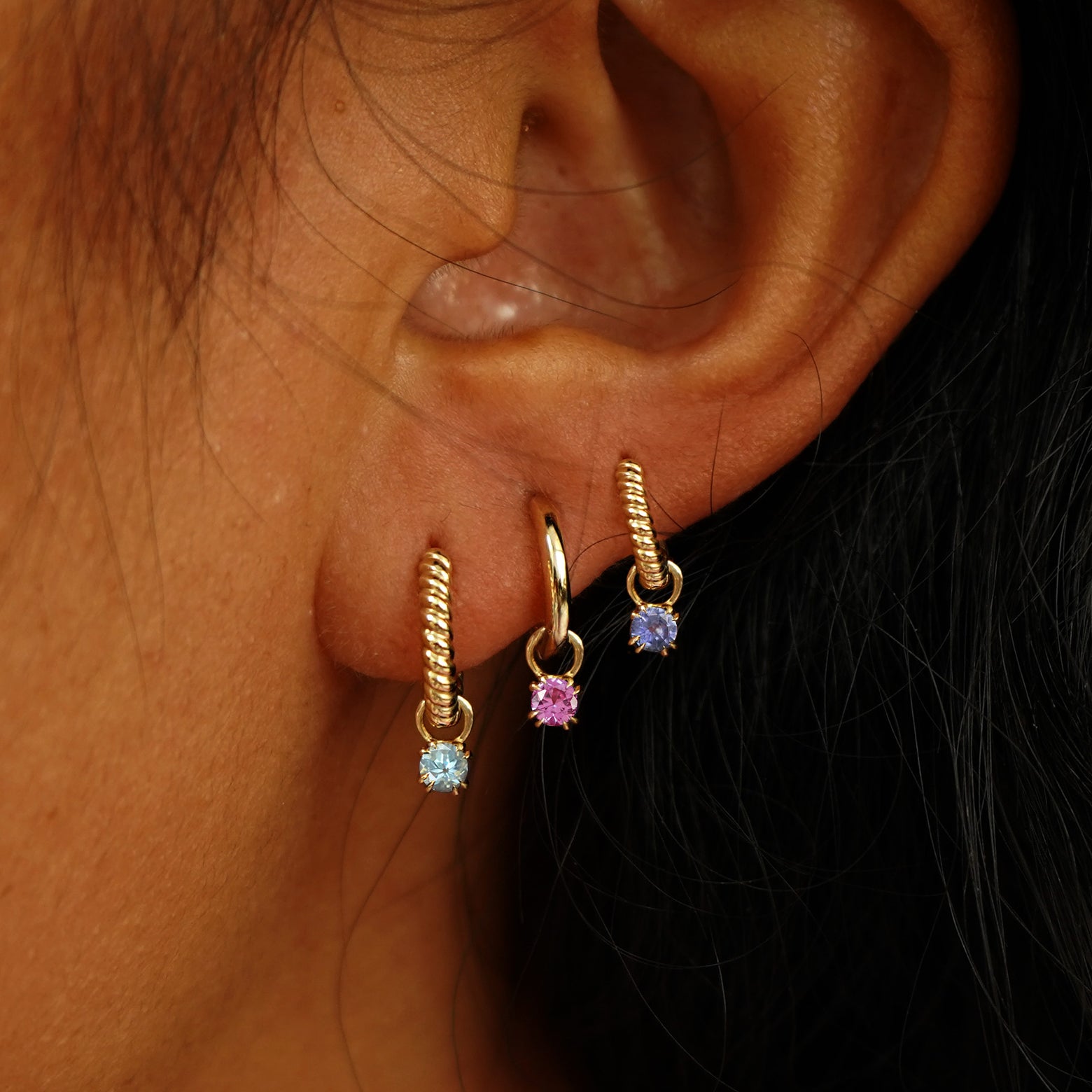 A model's ear wearing Aquamarine, Pink Sapphire, and Tanzanite charms on three different Huggie Hoop Earrings
