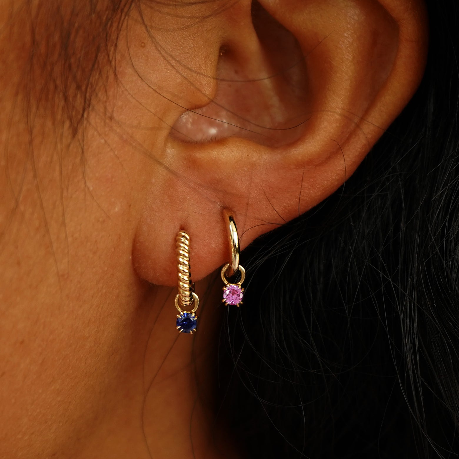 A model's ear wearing a Blue Sapphire Charm on a Rope Huggie and a Pink Sapphire Charm on a Curvy Huggie