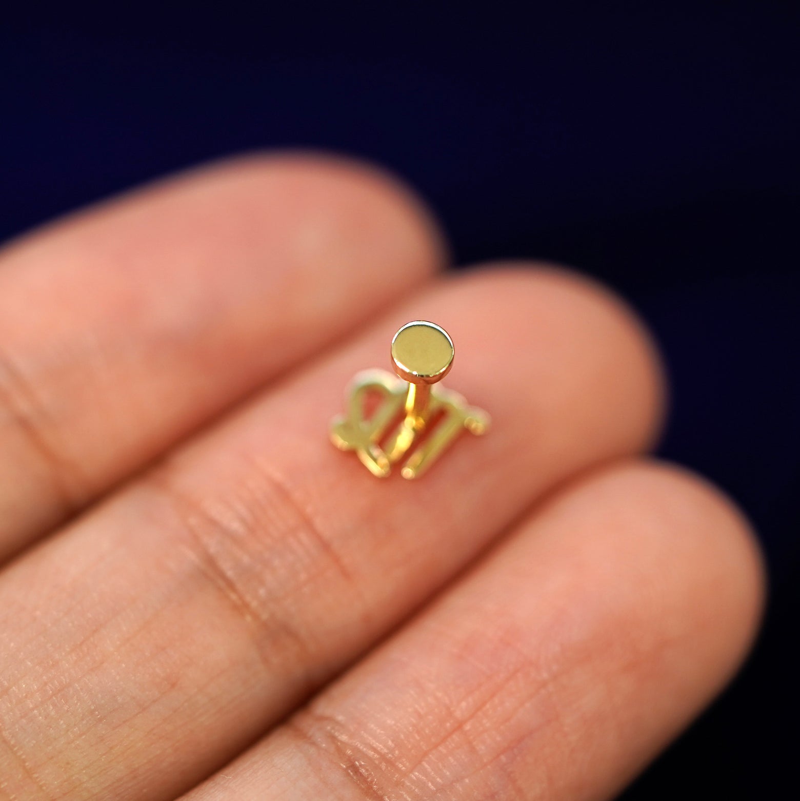 A solid yellow gold Virgo Horoscope Flatback Piercing laying facedown on a model's finger to show the end of the flat back