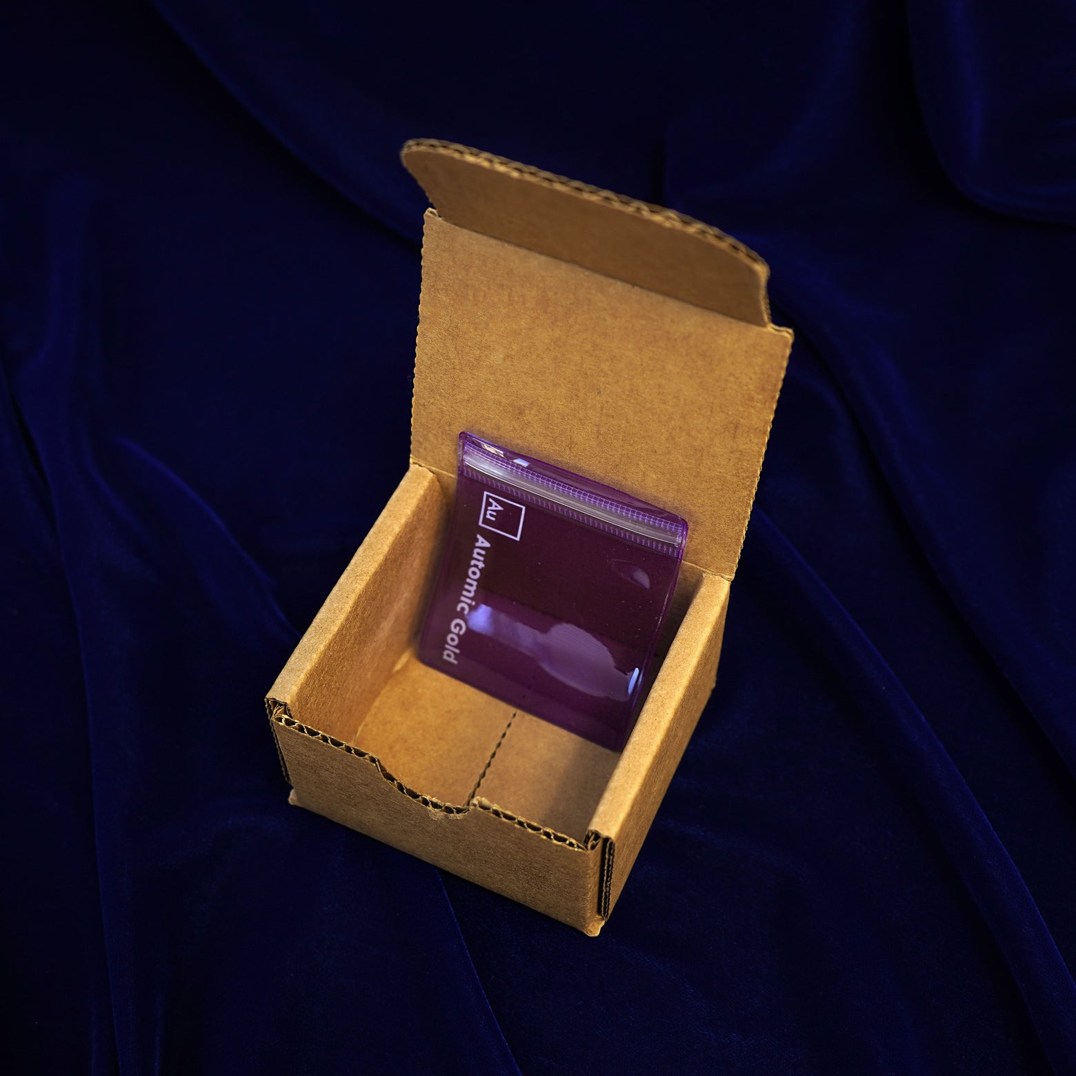 An open Automic Gold Minimal Packaging box on a dark blue background with a jewelry pouch inside