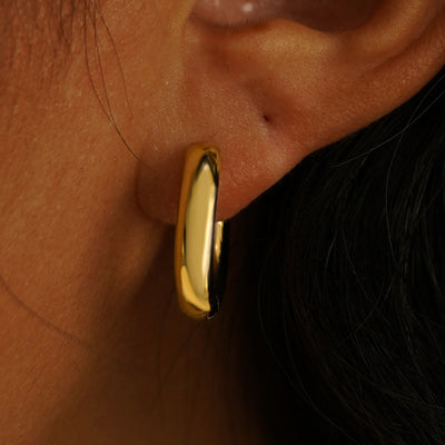 Close up view of a model's ear wearing a solid yellow gold Chunky Oval Huggie Hoop
