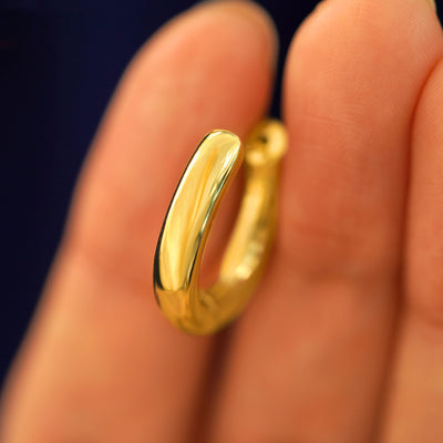 A 14k solid gold Chunky Oval Huggie Hoop between a model's fingers to show the thickness of the earring