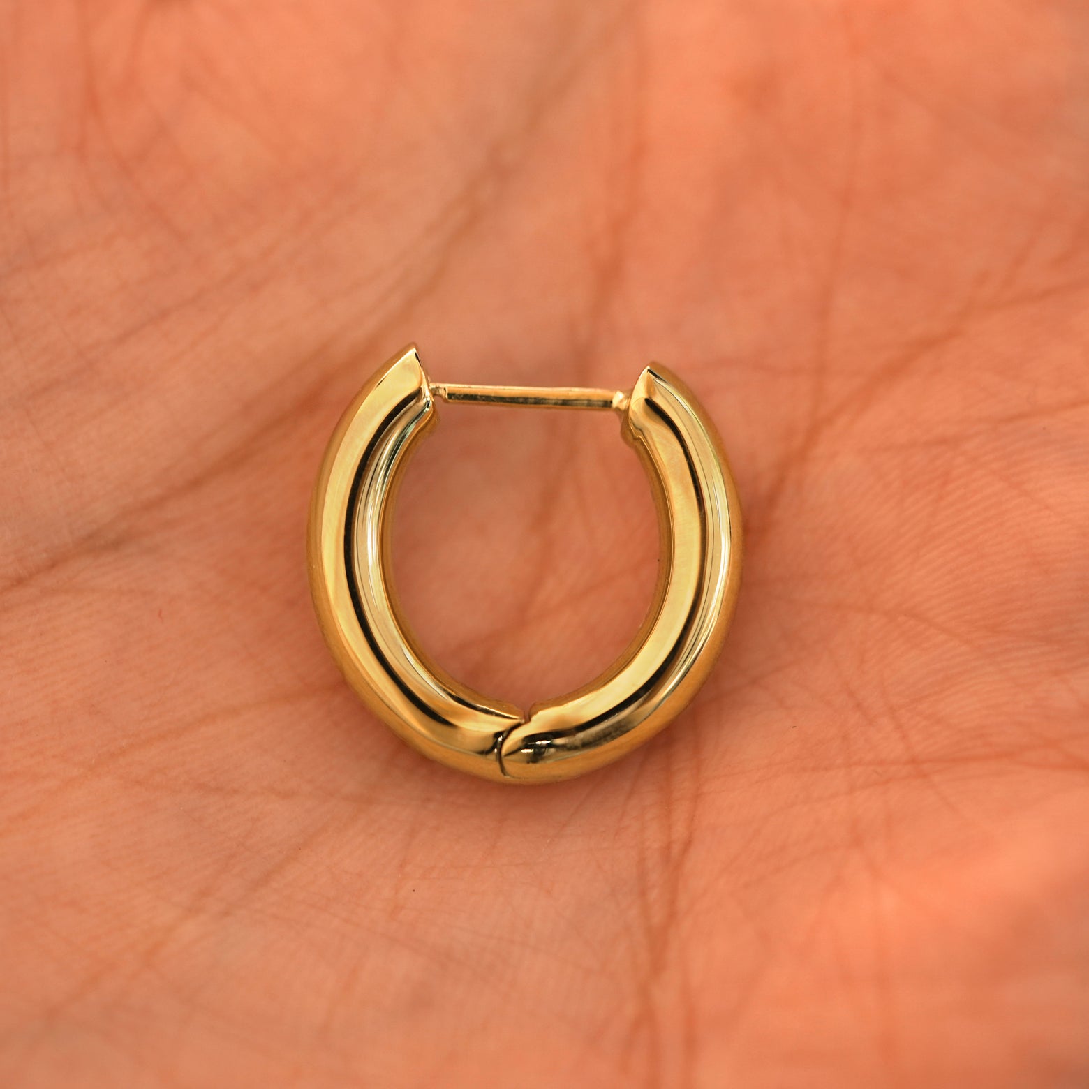 A closed single 14k yellow gold Chunky Oval Huggie Hoop resting in a model's palm