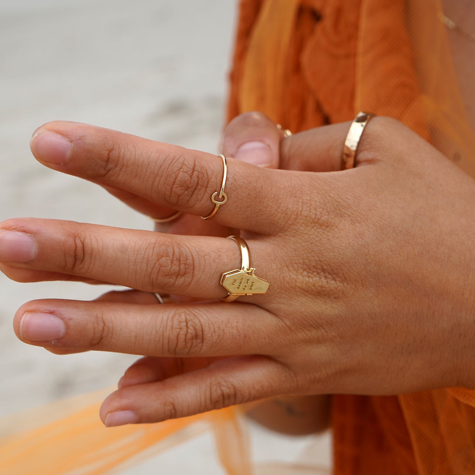 A model wearing orange clothing at the beach with their fingers steepled wearing several Automic Gold rings