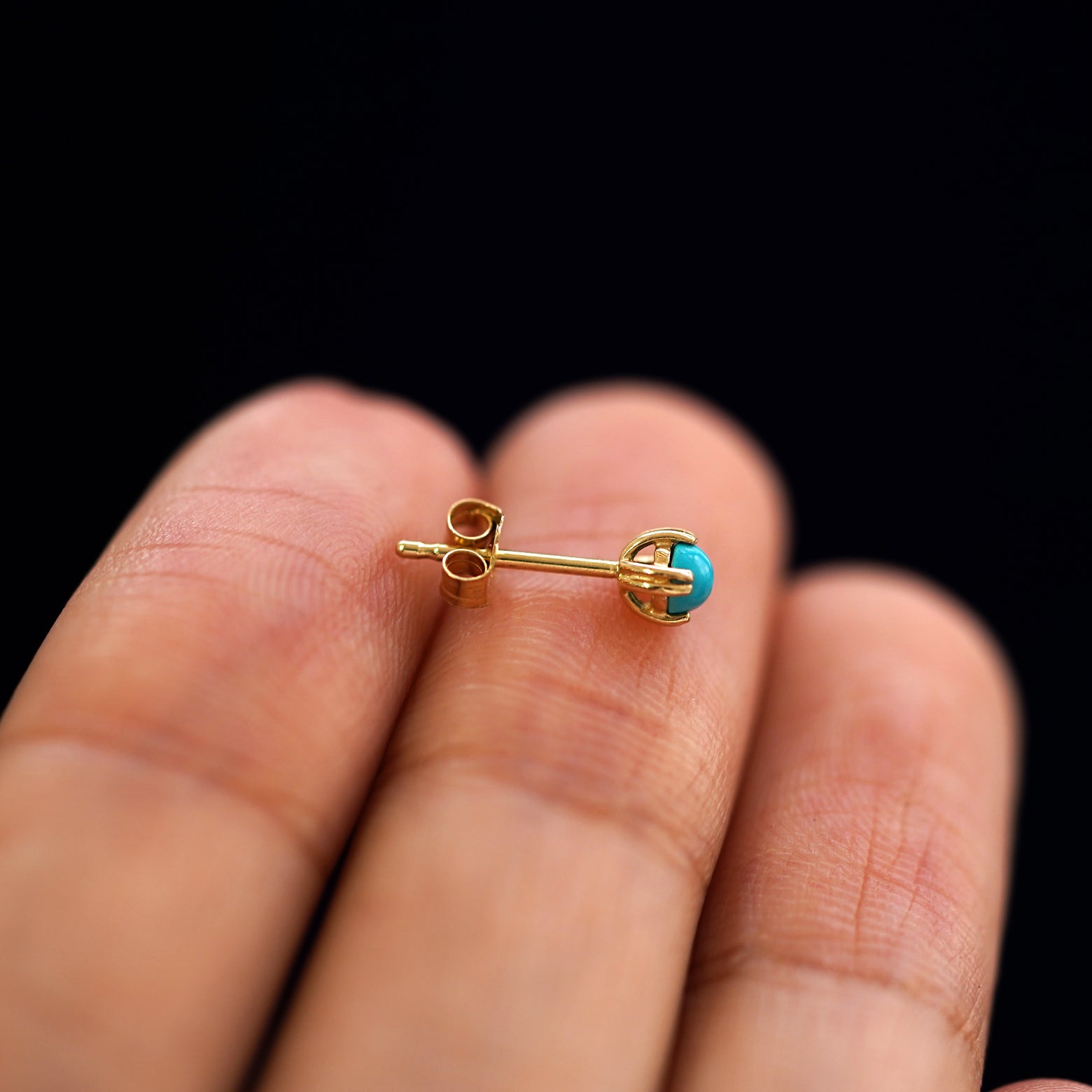 Side view of a 14k gold 3mm turquoise gemstone earring with a standard pushback backing on a model's finger