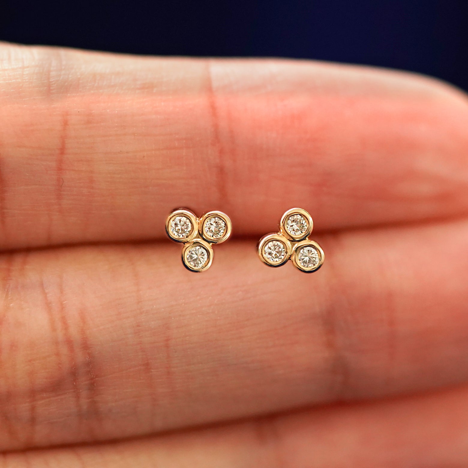 A hand holding a pair solid yellow gold Diamond Trio stud Flatback Piercings in between their fingers