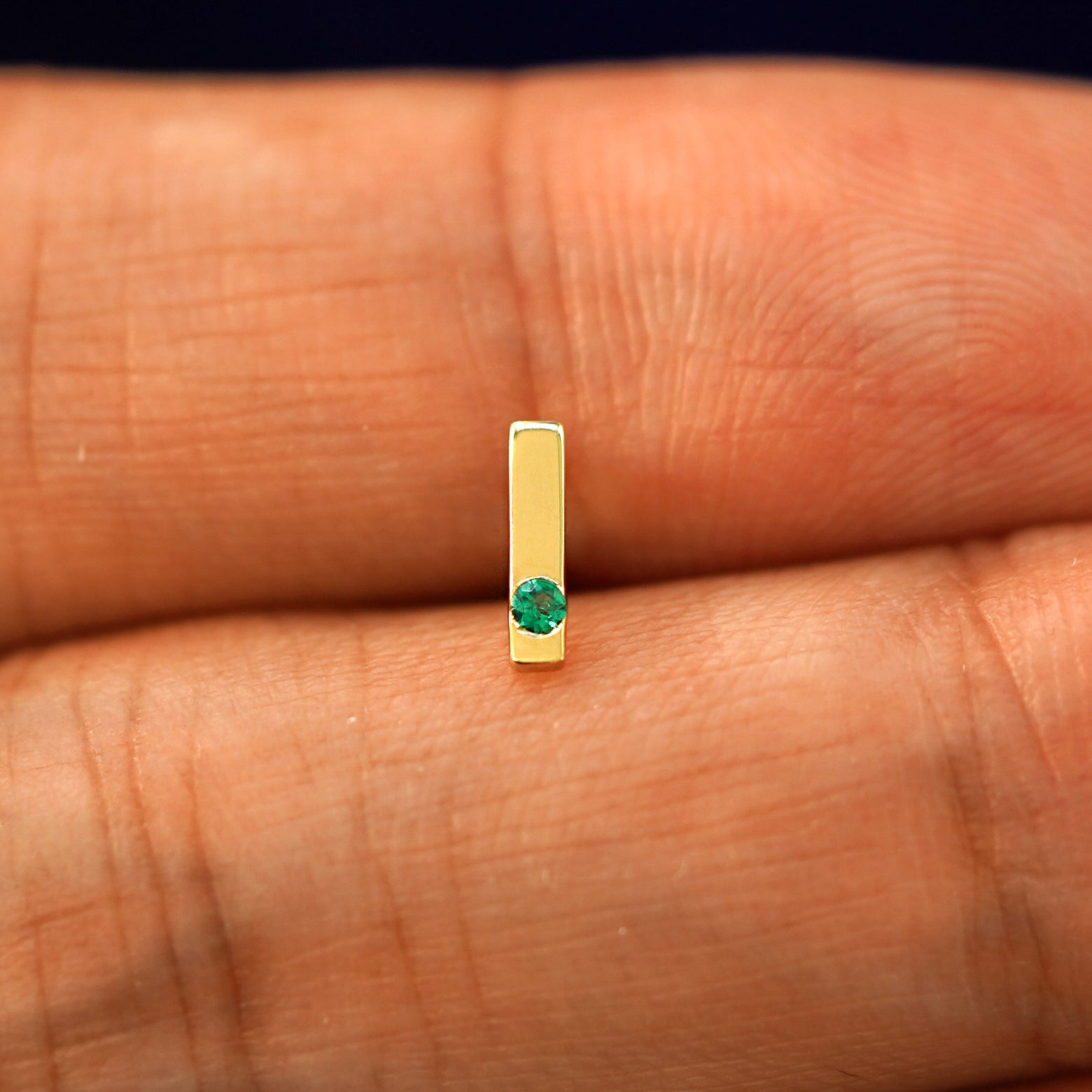 A model holding a single emerald Gemstone Bar Earring in yellow gold between two fingers