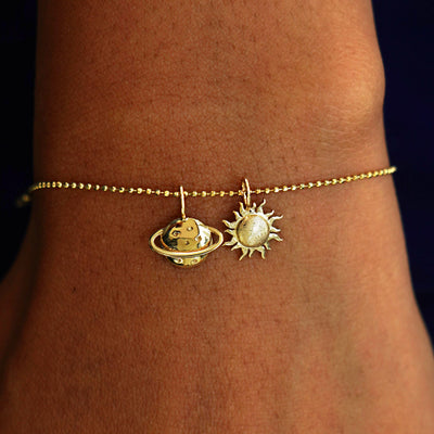 A model's wrist wearing a yellow gold Bead Chain with a Saturn Charm, and a Sun Charm