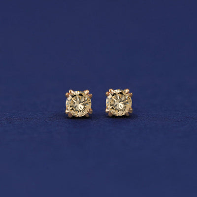 A pair of 14 karat gold stud earrings with 3 millimeter round Champagne diamonds on a dark blue background