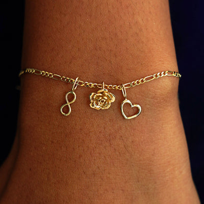 A model's wrist wearing a yellow gold Figaro Chain with an Inifnity Charm, a Rose Charm, and a Heart Charm