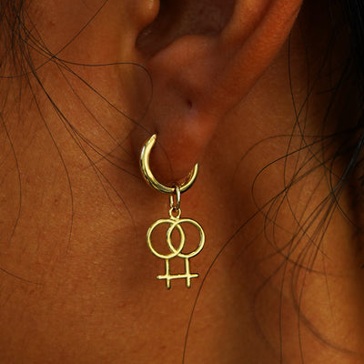 Close up view of a model's ear wearing a yellow gold Lesbian Symbol Charm on a Curvy Huggie Hoop