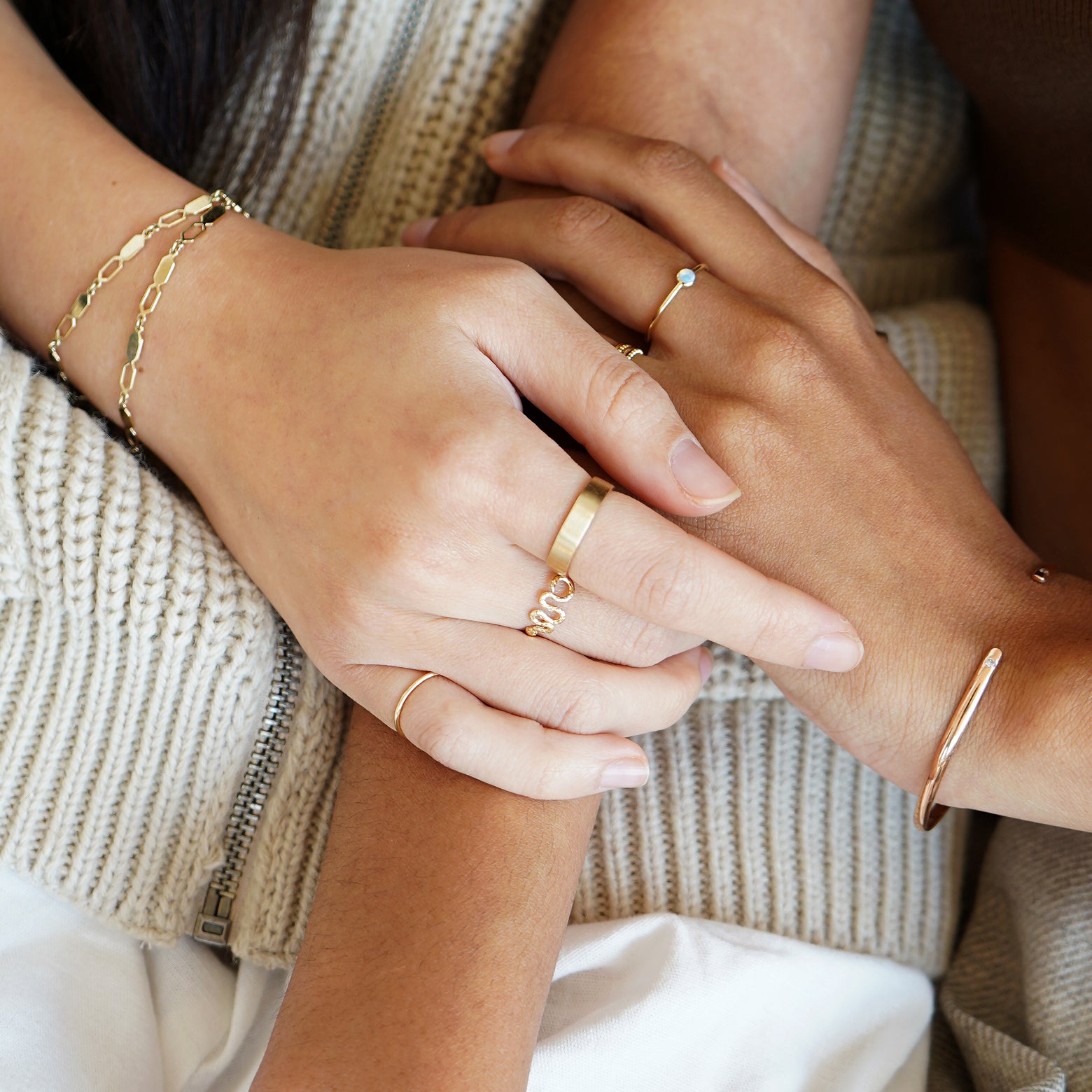 Two models touching each others hands over one model's arm while wearing various Automic Gold jewelry