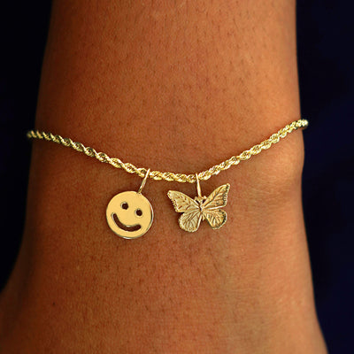 A model's wrist wearing a yellow gold Rope Chain with a Smiley Face Charm and a Butterfly Charm