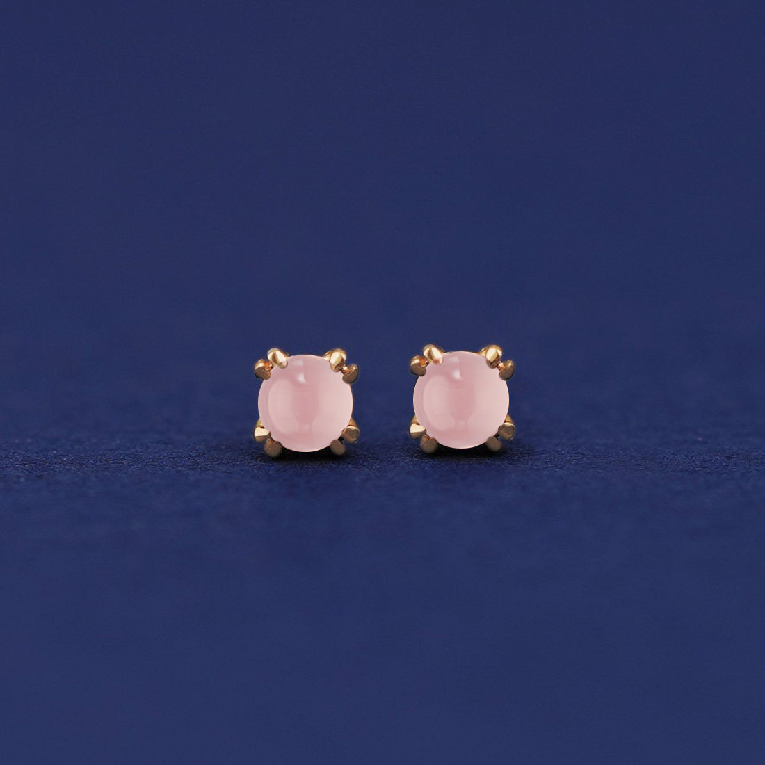 A pair of 14 karat gold studs earrings with 3 millimeter round Rose Quartz gemstones on a dark blue background