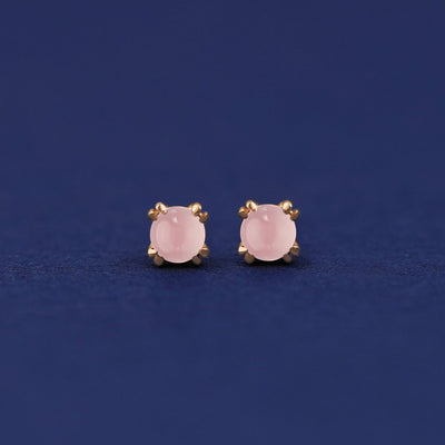 A pair of 14 karat gold studs earrings with 3 millimeter round Rose Quartz gemstones on a dark blue background