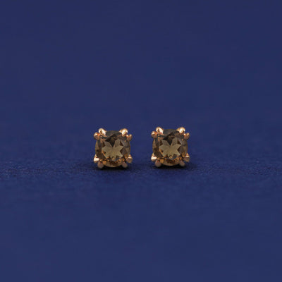 A pair of 14 karat gold studs earrings with 3 millimeter round Smoky Quartz gemstones on a dark blue background