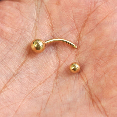 A yellow gold Large Curved Barbell Piercing with the externally threaded ball closure unscrewed in the palm of a model's hand