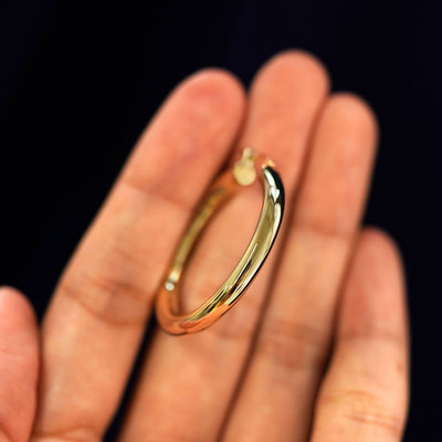 A 14k gold Large Tube Hoop Earring held in between a model's fingers to show the hoops thickness