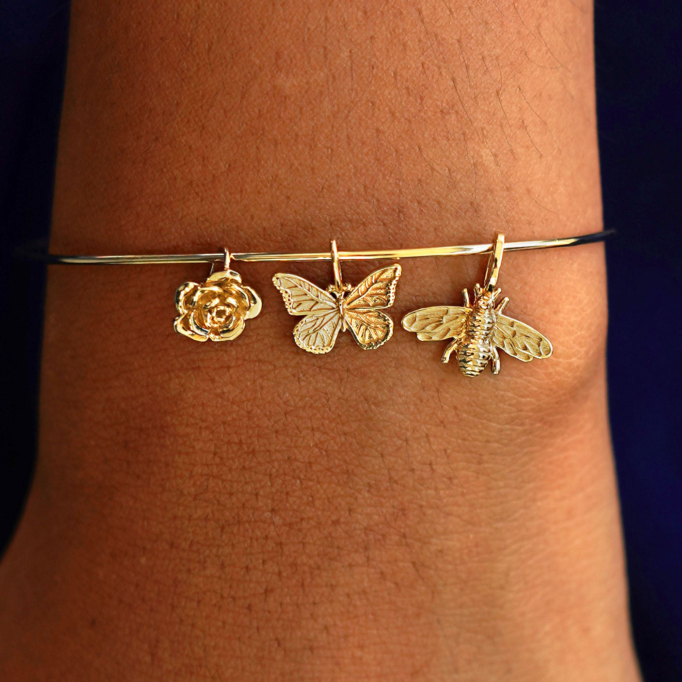 A model's wrist wearing a yellow gold Line Bangle with a Rose Charm, a Butterfly Charm, and a Bee Charm