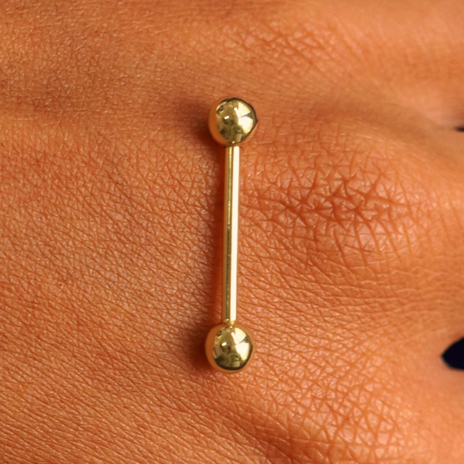 Close up view of a 14 karat gold Medium Straight Barbell Piercing resting on the back of a model's hand