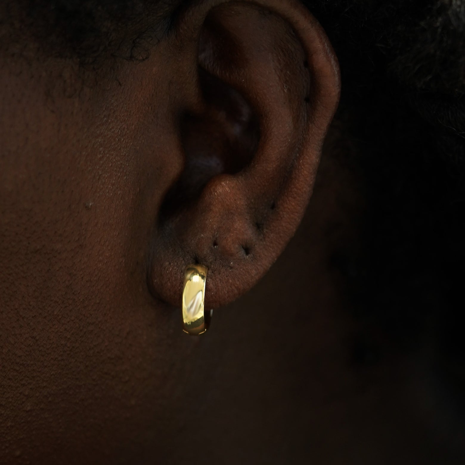 A model's ear wearing a 14k gold Medium Thick Huggie Hoop Earring