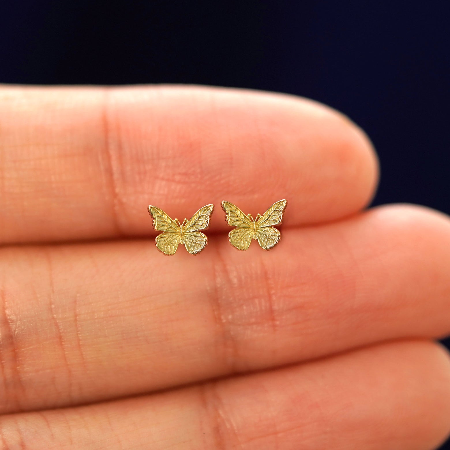 A hand holding a pair of solid yellow gold mini butterfly stud earrings in between their fingers