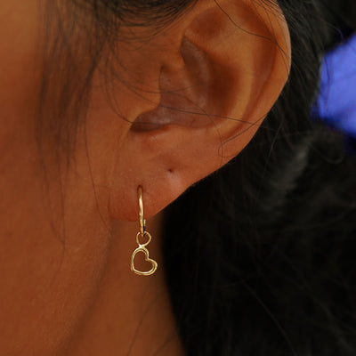 A model's ear wearing a 14k yellow gold Heart Charm on a Mini Seamless Huggie Earring