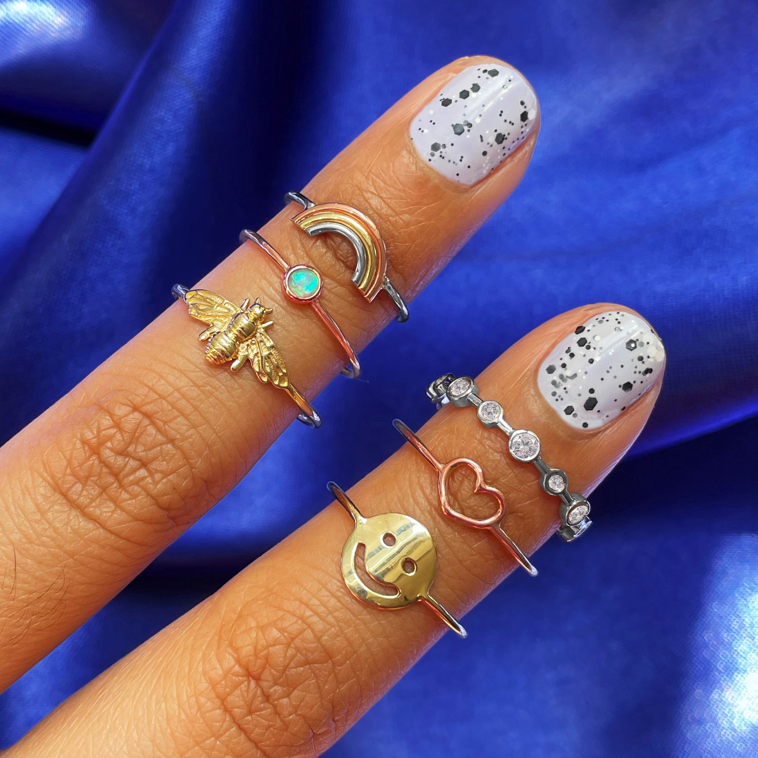 Two fingers with speckled nail polish wearing various Automic Gold rings including a rose gold Opal Ring