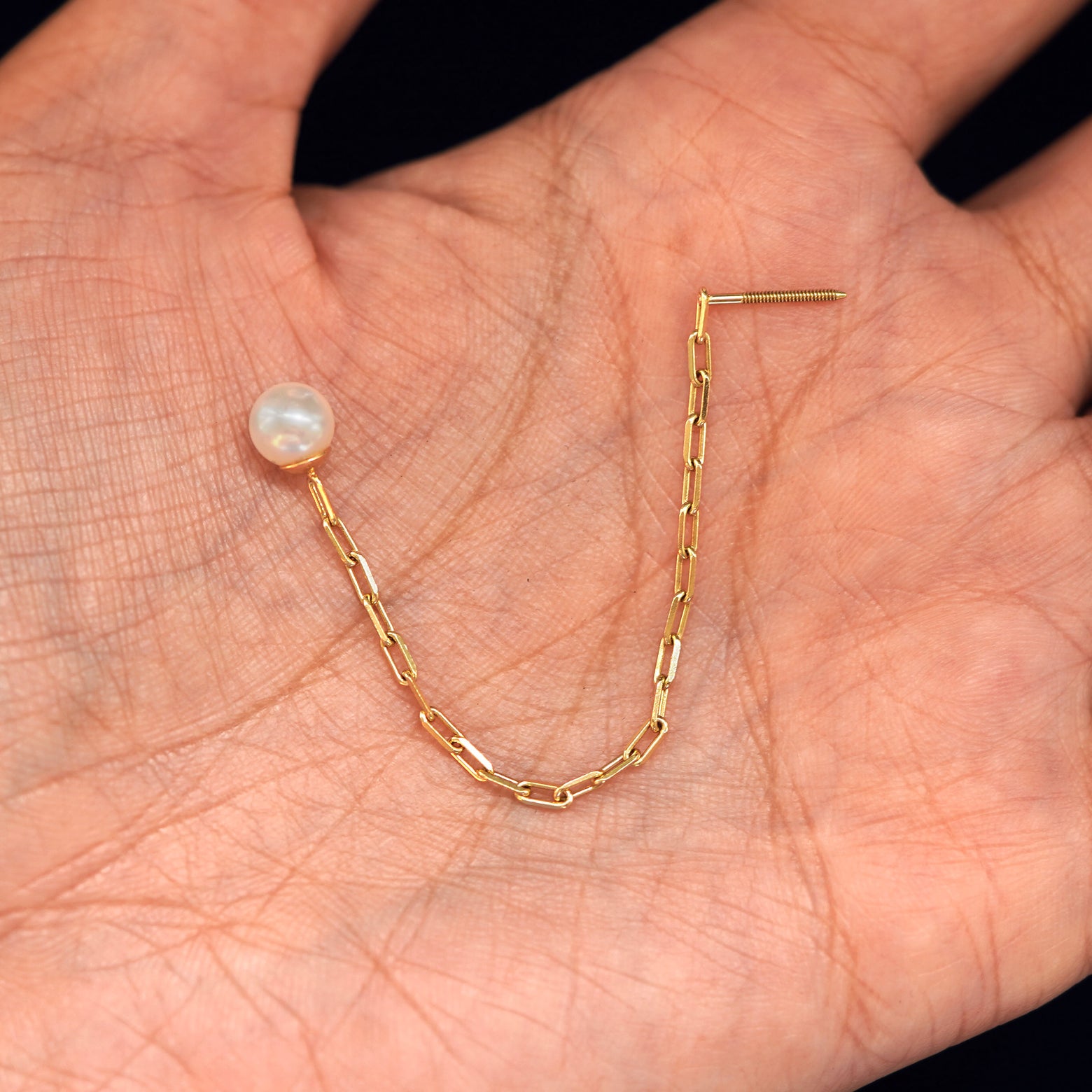 A solid gold Pearl Dangle Earring sitting in a model's palm with the butch chain in a arch and without its screwback backing