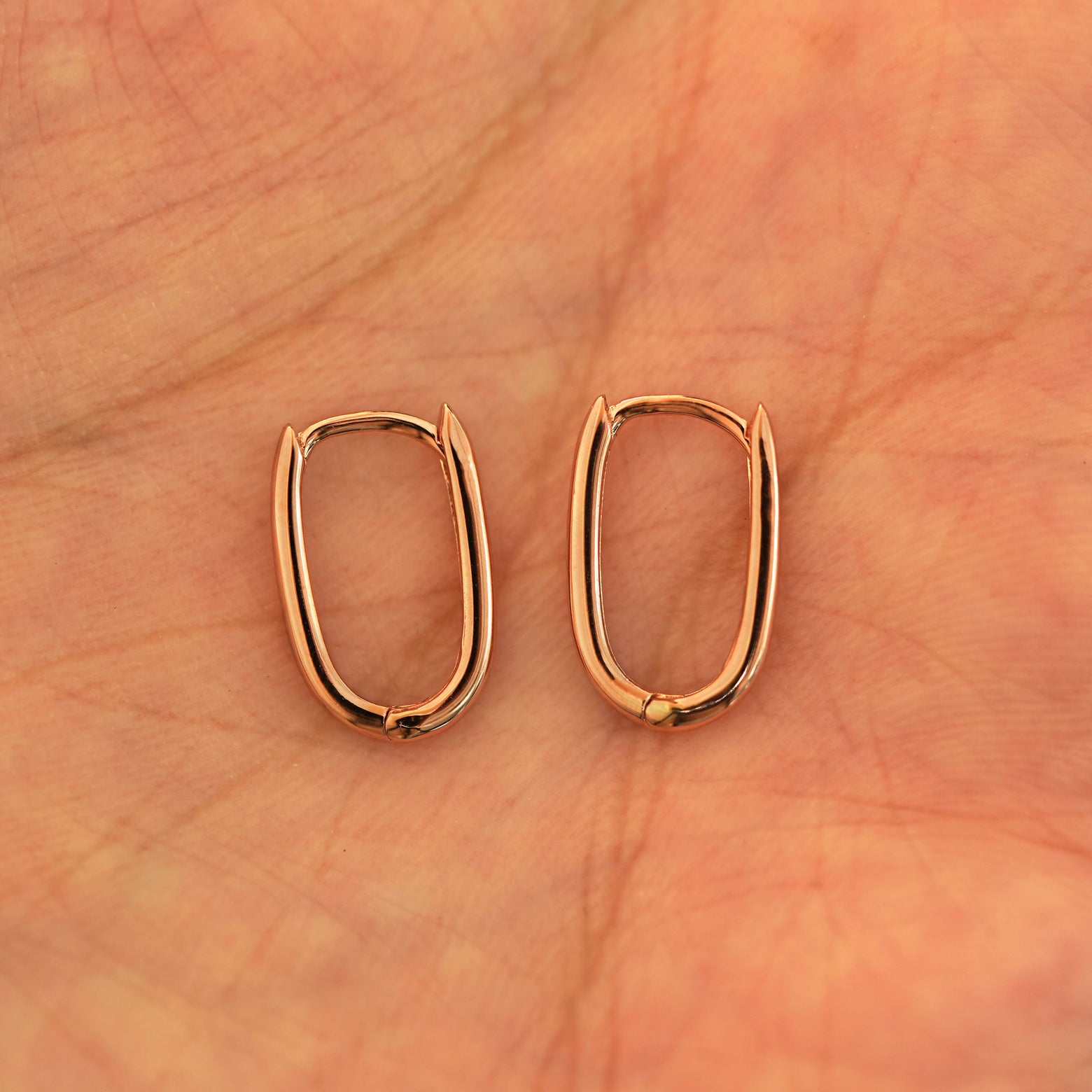 A pair of 14k rose gold Thin Oval Huggie Hoops resting in a model's palm