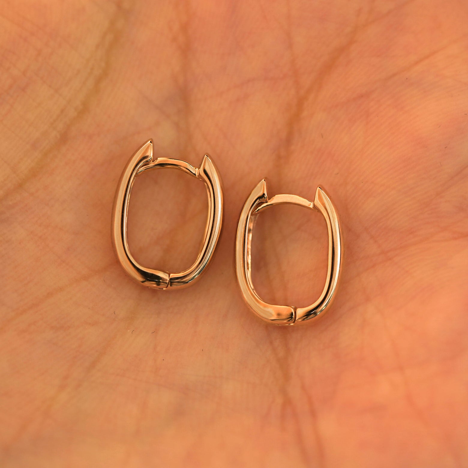 A pair of 14k rose gold Thick Oval Huggie Hoops resting in a model's palm
