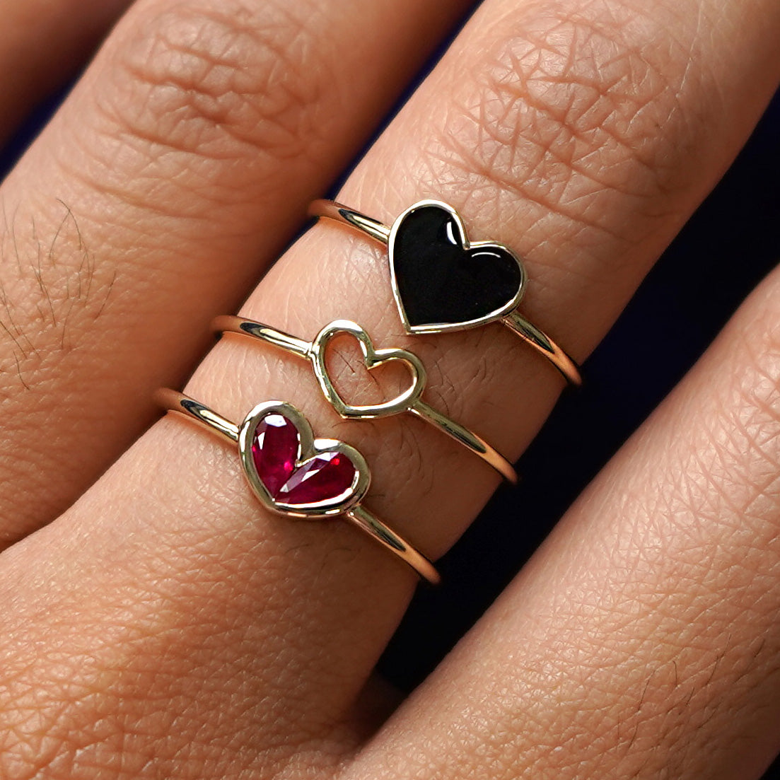 Close up view of a model's finger wearing a Ruby Heart Ring, a Heart Ring, and a black Enamel Heart Ring in a stack